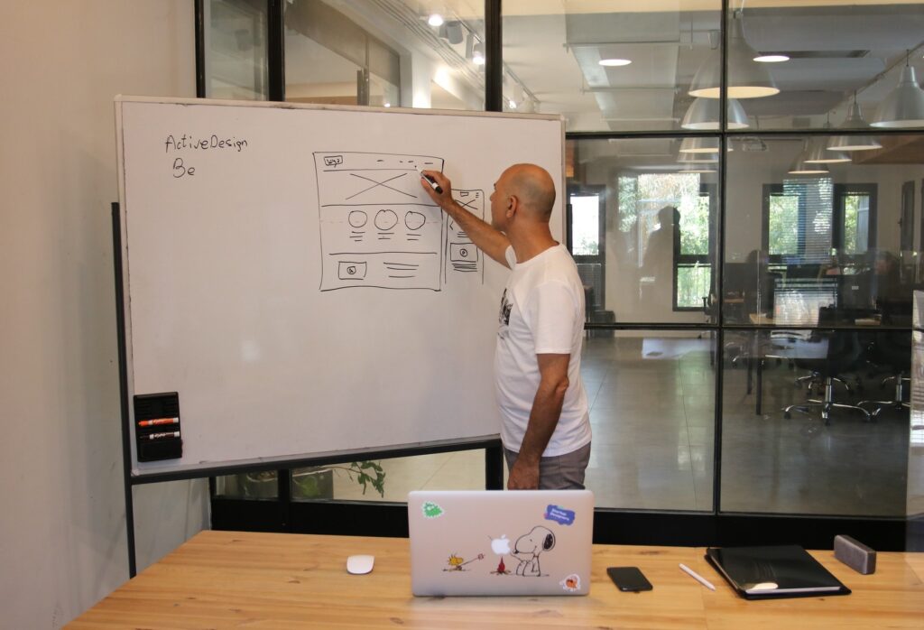 A white male UX designer is working on a wireframe on a whiteboard in an office.