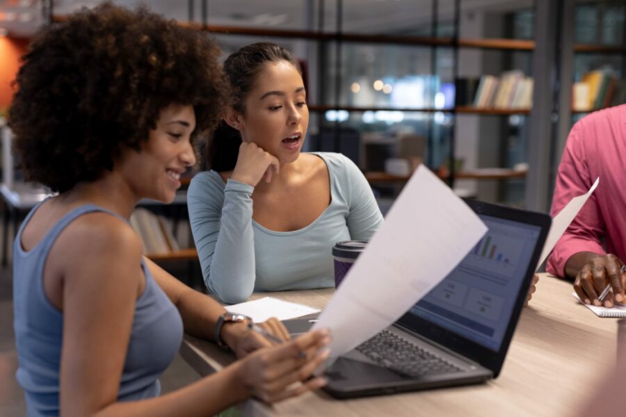 Two diverse women review files while seated at a table at work