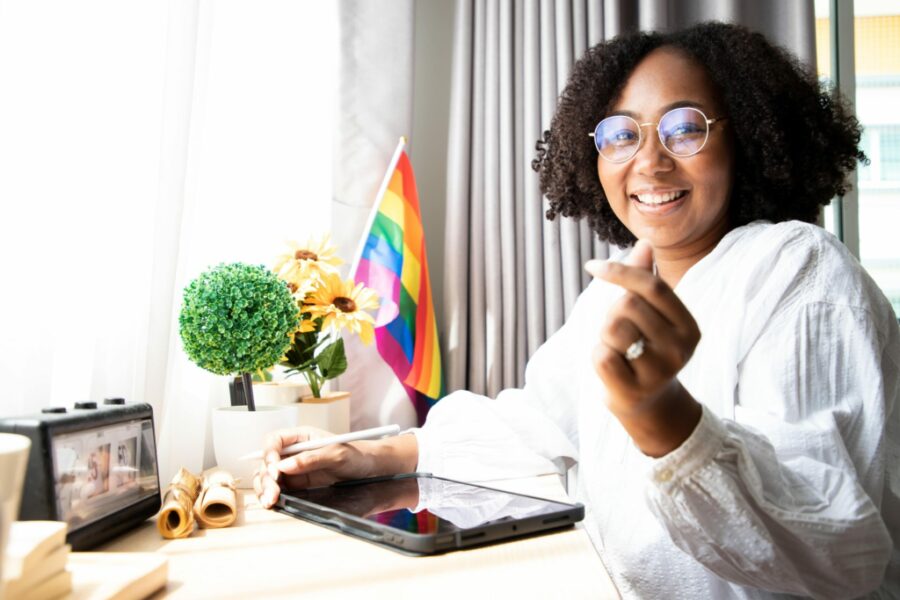 Woman works at her desk, looks at the camera and smiles, with a rainbow flag planted in a cup holder at her desk