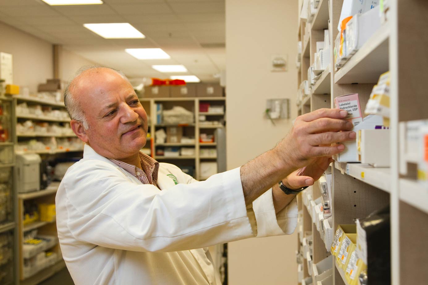 A pharmacist employed by a temp agency that's wearing a white lab coat and taking medicine off of a shelf.
