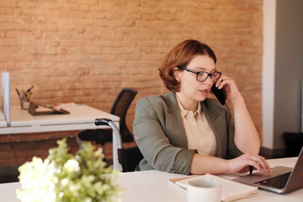 White woman sitting at desk, looking at laptop, talking on mobile phone